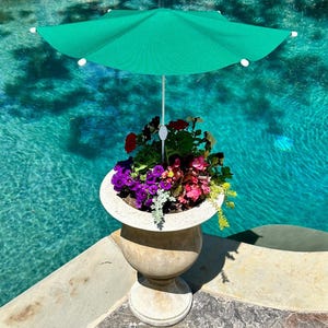 May include: A green patio umbrella with a white pole is mounted to a stone planter filled with colorful flowers. The planter is sitting on a stone patio next to a swimming pool.