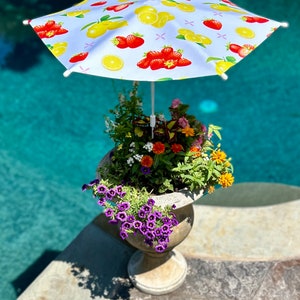 May include: A white patio umbrella with a lemon and strawberry print, positioned over a planter with purple, orange, and white flowers. The umbrella is positioned over a planter with a stone base.