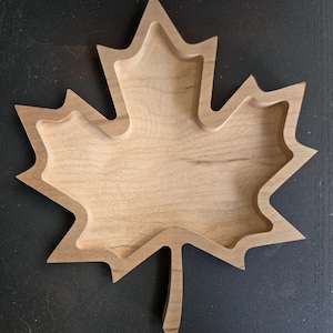 May include: A wooden maple leaf-shaped tray with a natural wood finish. The tray has a carved design with a raised edge and a central recessed area. The leaf's details are clearly defined, showcasing the wood's grain.