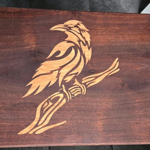 May include: A wooden cutting board with a detailed, laser-cut design of a raven perched on a branch. The raven is outlined in a light brown color, contrasting with the dark brown wood of the board.