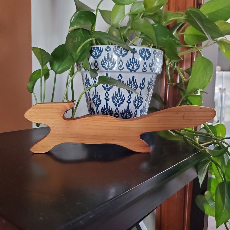 K&ouml;nnte beinhalten: Eine Holzfigur eines Hasen in einer Sprungpose, geschnitzt aus hellem braunem Holz. Die Figur steht auf einer dunkelbraunen Oberfl&auml;che, vor einer Topfpflanze mit gr&uuml;nen Bl&auml;ttern und einem wei&szlig;en Keramiktopf mit blau-wei&szlig;em Muster.
