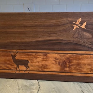 Könnte beinhalten: Rechteckiges Holzschneidebrett mit einer Hirschsilhouette und zwei fliegenden Gänsen. Das Brett besteht aus dunkelbraunem und hellbraunem Holz mit einem natürlichen Holzmaserungsmuster. Der Hirsch befindet sich im unteren Bereich und die Gänse im oberen Bereich.