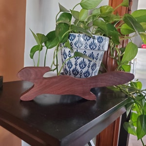 K&ouml;nnte beinhalten: Eine Holzskulptur eines stilisierten Kaninchens, in dunkelbraun lackiert, steht auf einem schwarzen Tisch. Im Hintergrund befindet sich eine Topfpflanze mit gr&uuml;nen Bl&auml;ttern und einem wei&szlig;-blau gemusterten Topf.