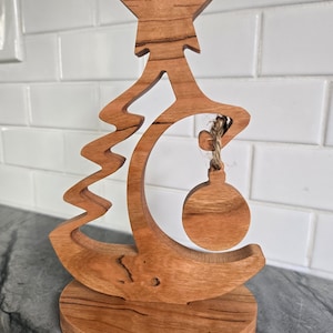 Op de afbeelding: Een houten kerstboom-versieringhouder met een ster bovenop en een ronde uitsparing voor een ornament. De houder is gemaakt van lichtbruin hout met een natuurlijke houtnerf.