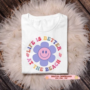 May include: White t-shirt with a colorful graphic that reads "LIFE IS BETTER AT THE BEACH" around a smiling flower. The design includes a digital download icon. The shirt is folded on a fluffy, white surface.