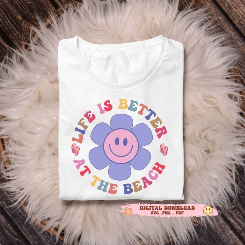 May include: White t-shirt with a colorful graphic that reads "LIFE IS BETTER AT THE BEACH" around a smiling flower. The design includes a digital download icon. The shirt is folded on a fluffy, white surface.