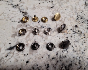 May include: Assortment of metal snap fasteners in various finishes, including silver, gold, and black. The snaps are arranged on a light gray speckled surface.