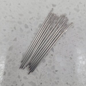 May include: A bundle of silver sewing needles, arranged in a fan shape on a white speckled surface.