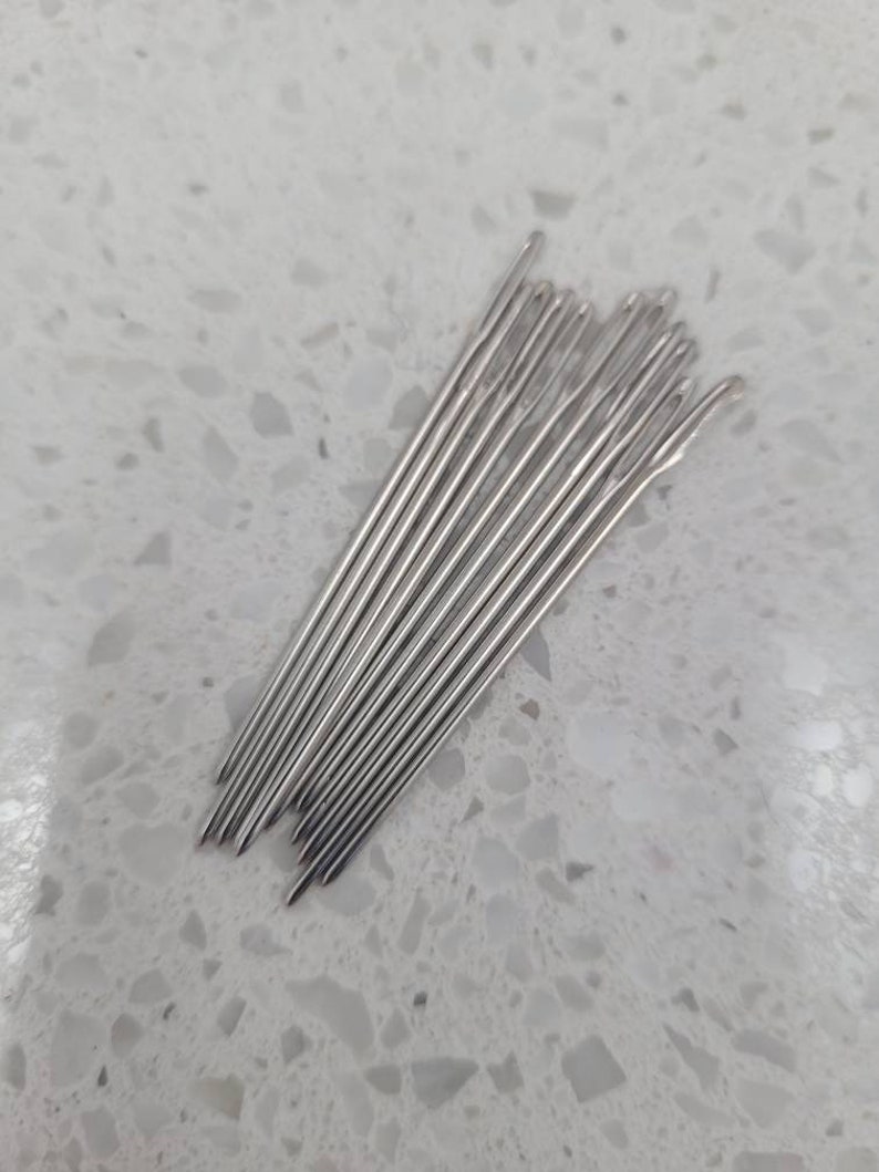 May include: A bundle of silver sewing needles, arranged in a fan shape on a white speckled surface.