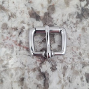 May include: A silver metal buckle with a smooth finish. The buckle has two loops and a center bar.