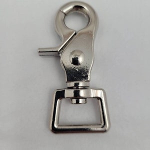 May include: A silver-colored metal swivel snap hook. The hook has a spring-loaded clasp and a rotating eyelet. The base has a rectangular loop. The hardware is designed for attaching to a leash or strap.