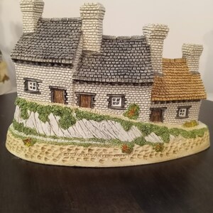 Pode incluir: Estatueta detalhada em miniatura de uma casa de campo com três chaminés, telhados de telhas cinzentas e paredes de tijolo branco. A base apresenta um muro de pedra com folhagem verde e uma base bege texturizada. A estatueta tem pequenas janelas e portas castanhas.