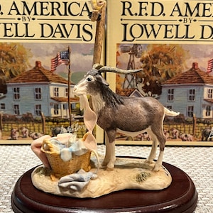 May include: A brown and white goat figurine with a basket of laundry next to it. The figurine is on a wooden base with a brown and white patterned surface. The figurine is part of a collection called "R.F.D. America" by Lowell Davis.