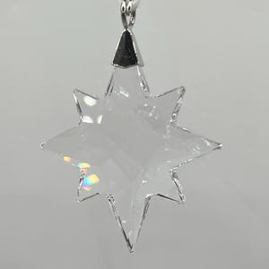 Puede incluir: Adorno transparente en forma de estrella facetada con una tapa de metal plateado y una cuerda blanca para colgar. La estrella tiene ocho puntas y refleja un arcoíris de colores. Un artículo decorativo para las fiestas o todo el año.