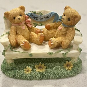 May include: A decorative figurine featuring two teddy bears seated on a bench. The bears are light brown with pink and blue ribbons, and a heart on their chests. The bench has a banner that reads "Heart to Heart". The base is green with flowers.