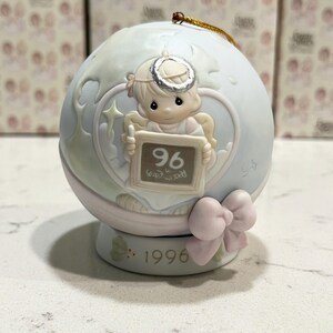 May include: A porcelain Christmas ornament featuring a Precious Moments angel holding a chalkboard with the number 96 on it. The ornament is blue and white with a pink bow and the year 1996 on the base.