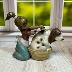 Puede incluir: Una figurita de cerámica de una niña cepillando a un perro. La niña lleva un vestido marrón y una falda azul. El perro es marrón y blanco y está sentado en una bañera de cerámica marrón.