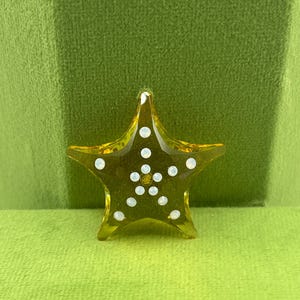 Puede incluir: Un objeto decorativo en forma de estrella amarilla con adornos circulares blancos. La estrella está hecha de un material translúcido y se encuentra sobre un fondo verde. El objeto tiene cinco puntas y una superficie ligeramente texturizada.