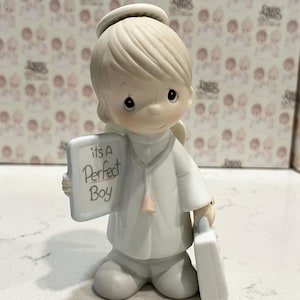 May include: A Precious Moments figurine of an angel holding a sign that reads "It's A Perfect Boy." The angel is wearing a white robe and has a halo. It also carries a white briefcase. The figurine is a light cream color.