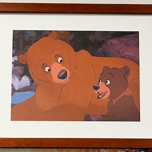 May include: A framed print featuring an animated illustration of two bears. The larger bear is brown with a light brown muzzle, and the smaller bear is dark brown. The background includes a forest setting.