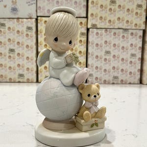 May include: A porcelain figurine of a cherub angel with blonde hair and a halo, sitting on a globe with a small teddy bear wearing a pink bow sitting on a box beside the globe. The figurine is white with blue accents.