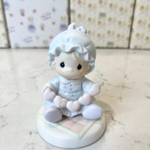 以下が含まれることがあります： 水色の赤ちゃんの置物。帽子をかぶり、ピンクのハートのガーランドを持っています。丸い白い台座に座っており、ピンクと白の模様があります。背景には同様のデザインの箱があります。