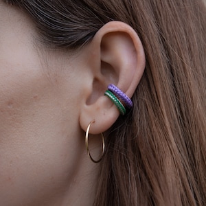 Könnte beinhalten: Eine Person trägt einen goldenen Creolenohrring und zwei Ohrklemmen, eine grüne und eine violette.