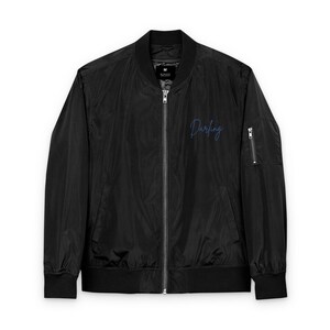 May include: Black bomber jacket with a zipper closure, two front pockets, and a small zippered pocket on the left sleeve. The word "Darling" is written in cursive script on the left chest.