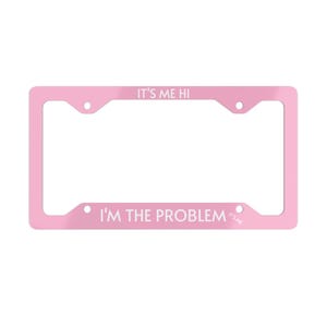 以下が含まれることがあります： 白い文字で「It's Me Hi I'm The Problem It's Me」と書かれたピンクの金属製ナンバープレートフレーム