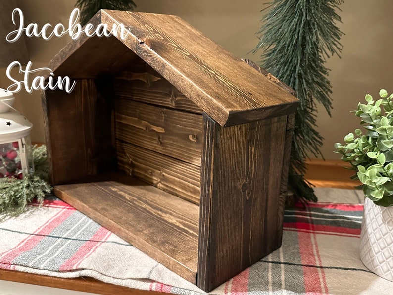 Peut inclure: Une cr&egrave;che en bois avec une finition Jacobean fonc&eacute;e. La structure a un toit en pente et une base rectangulaire. Le texte "Jacobean Stain" est visible. D&eacute;coration de No&euml;l pour une sc&egrave;ne de la Nativit&eacute;.