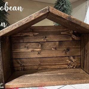 Peut inclure: Une cr&egrave;che en bois de couleur marron fonc&eacute; avec un toit en pente. La structure est faite de planches de bois horizontales. Le texte "Jacobean Stain" est visible en haut &agrave; gauche. Le design est rustique et artisanal.