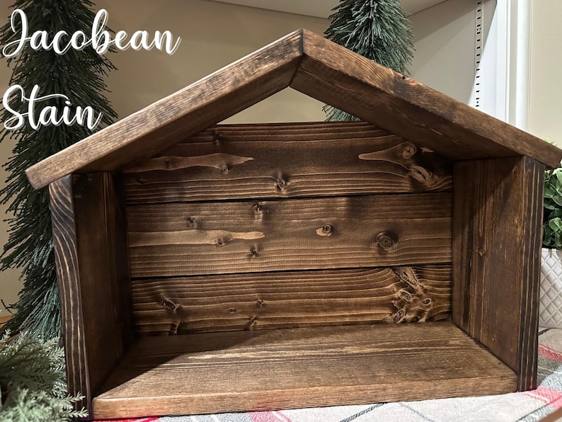 Peut inclure: Une cr&egrave;che en bois de couleur marron fonc&eacute; avec un toit en pente. La structure est faite de planches de bois horizontales. Le texte "Jacobean Stain" est visible en haut &agrave; gauche. Le design est rustique et artisanal.