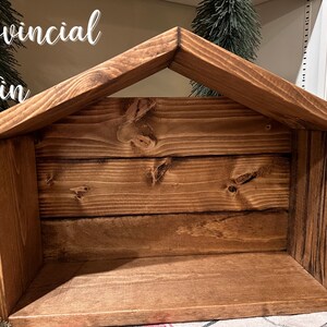 Peut inclure: Une cr&egrave;che en bois avec une finition de teinture provinciale. La structure est faite de planches et pr&eacute;sente un toit en pente. Le texte "Provincial Stain" est visible en haut &agrave; gauche. La cr&egrave;che est con&ccedil;ue pour contenir des figurines.