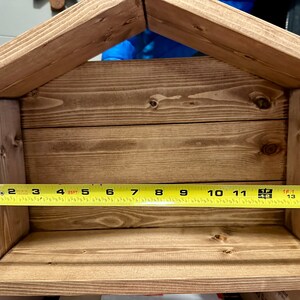 Peut inclure: Une &eacute;tag&egrave;re en bois en forme de nichoir, teinte dans une teinte marron chaud. L'&eacute;tag&egrave;re mesure environ 35,5 cm de large, avec un dessus en forme de toit. La construction pr&eacute;sente un grain de bois visible et un design simple et rustique.
