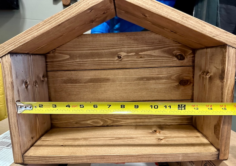 Peut inclure: Une &eacute;tag&egrave;re en bois en forme de nichoir, teinte dans une teinte marron chaud. L'&eacute;tag&egrave;re mesure environ 35,5 cm de large, avec un dessus en forme de toit. La construction pr&eacute;sente un grain de bois visible et un design simple et rustique.
