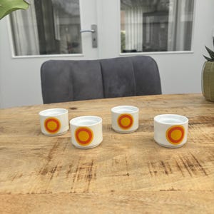 Op de afbeelding: Vier witte keramische eierdopjes met een retro design. Elke beker heeft een centrale oranje cirkel omgeven door een gele ring. De bekers staan op een houten tafel.