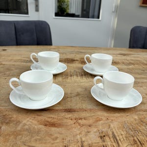 Op de afbeelding: Vier witte porseleinen theekopjes en schotels op een houten tafel. Elk kopje heeft een gebogen oor en staat op een bijpassende schotel. De scène is helder verlicht, met natuurlijk licht.