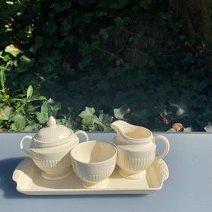 Peut inclure: Un service à thé crème sur un plateau rectangulaire. L'ensemble comprend un sucrier avec couvercle, un petit bol et une crémière. Le plateau et les pièces ont un design côtelé. Le fond présente un feuillage vert et une surface bleue.