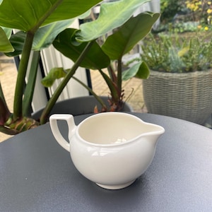Può includere: Un brocca per la panna in ceramica bianca con un corpo arrotondato e un piccolo beccuccio. La brocca ha un manico sul lato sinistro e si trova su una superficie grigio scuro.