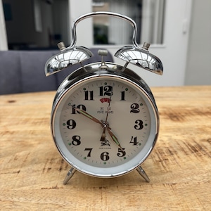 Op de afbeelding: Een zilverkleurige retro-wekker met een witte wijzerplaat en zwarte cijfers. De klok heeft twee bellen bovenop en drie kleine pootjes. De wijzerplaat toont de tekst "FIVE RAMS" en "MADE IN CHINA".