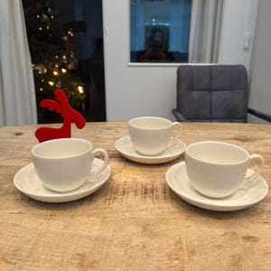 Könnte beinhalten: Drei weiße Keramik-Teetassen und Untertassen stehen auf einem hellbraunen Holztisch. Eine rote Rentierdekoration befindet sich auf dem Tisch. Der Hintergrund umfasst ein Fenster und einen Weihnachtsbaum.