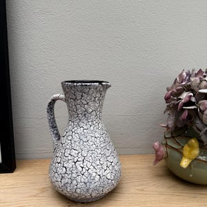 Op de afbeelding: Een keramische kan met een getextureerde craquelé glazuur in wit en grijs. De kan heeft een handvat en een donkere rand. Het staat op een houten oppervlak, met een glimp van een omlijst object en een decoratieve vaas met bloemen.