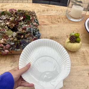 Op de afbeelding: Een witte, schelpvormige serveerschaal met een geribbeld ontwerp. De schaal wordt vastgehouden door een persoon die een blauwe longsleeve draagt. De schaal staat op een houten tafel, met een bloemstuk en een kleine pompoendecoratie.