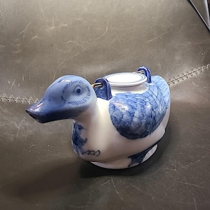 May include: A white and blue ceramic duck teapot. The teapot has a detailed blue pattern on the wings and head, with a small, round lid. The spout and handle are also blue, creating a decorative piece for serving tea.