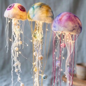 May include: Three hanging jellyfish decorations crafted from felted wool in cream, yellow, blue, and purple hues. Each jellyfish features long, dangling tentacles adorned with white beads and thin, glowing LED string lights for illumination.