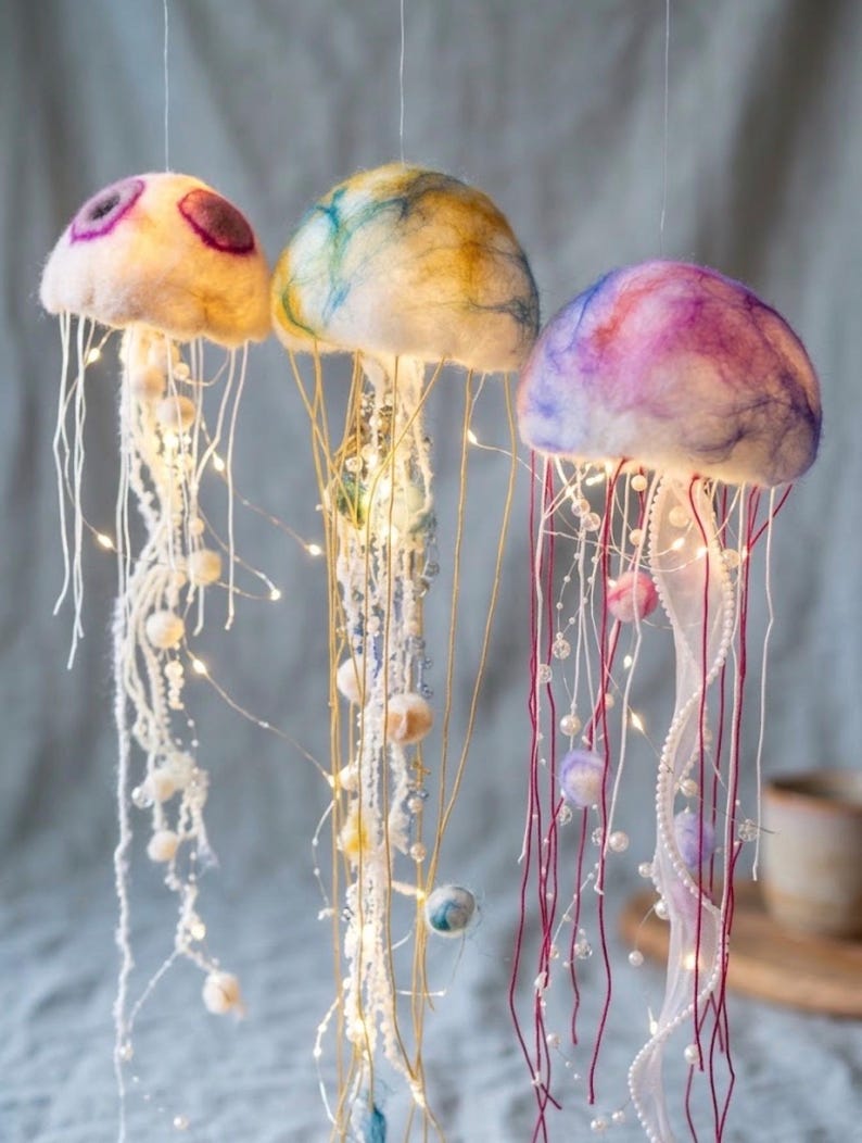 May include: Three hanging jellyfish decorations crafted from felted wool in cream, yellow, blue, and purple hues. Each jellyfish features long, dangling tentacles adorned with white beads and thin, glowing LED string lights for illumination.