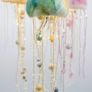 May include: Three hanging jellyfish sculptures crafted from felted wool. The left one is cream with pink spots, the center is a blend of green and blue, and the right is purple and pink. Each features long, thin dangling tentacles with small beads.