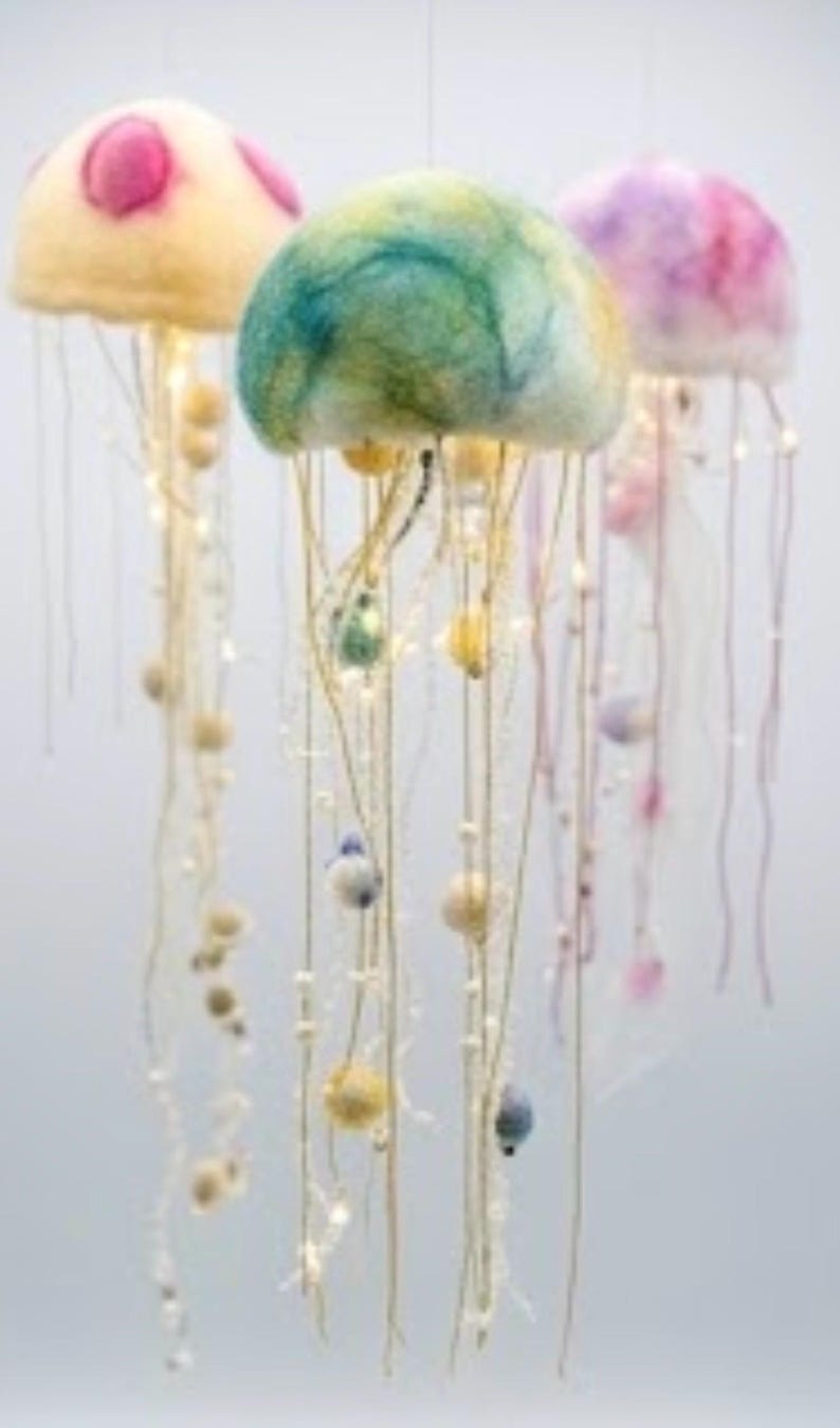 May include: Three hanging jellyfish sculptures crafted from felted wool. The left one is cream with pink spots, the center is a blend of green and blue, and the right is purple and pink. Each features long, thin dangling tentacles with small beads.