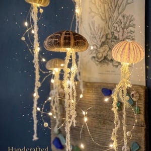 May include: Handmade luminous reef art showcasing jellyfish-shaped lights with warm white LED lights. The jellyfish are crafted from natural materials and suspended from above. The background includes a wooden plank with sea glass and a vintage coral print.