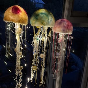 May include: Three hanging jellyfish decorations crafted from felted wool in yellow, green, and pink hues. Each jellyfish features long, dangling tentacles adorned with small beads and integrated warm white LED fairy lights that illuminate the structures.